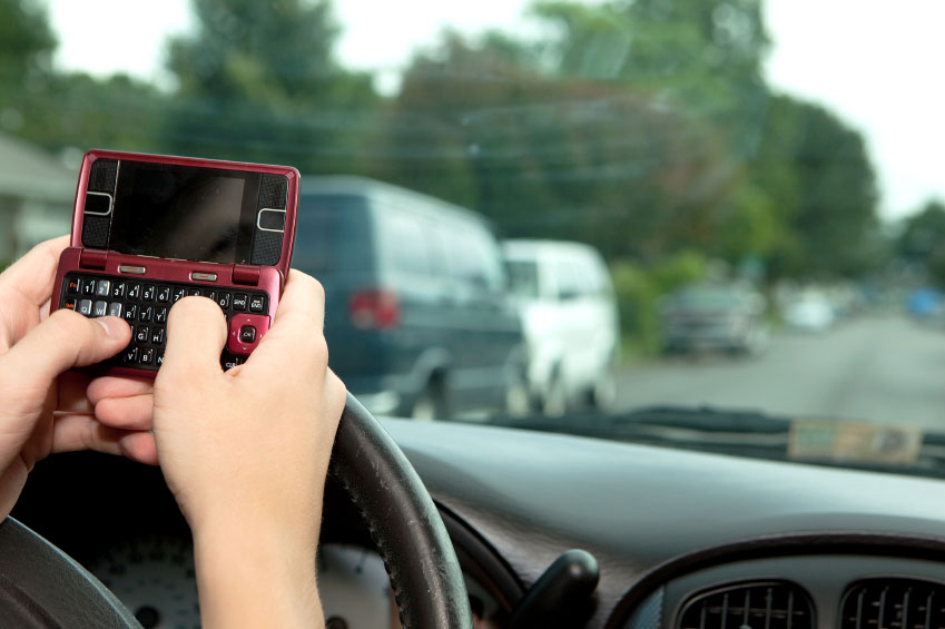 Hands holding up a pink phone to their person behind the steering wheel of a car.