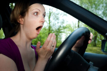 Woman wearing a purple shirt acting surprised by holding her mouth open behind the wheel of a car.