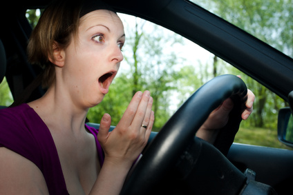 Woman wearing a purple shirt acting surprised by holding her mouth open behind the wheel of a car.