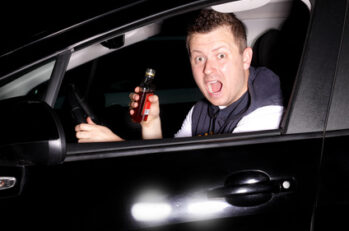 Guy acting surprised as he gets caught drinking liquor behind the wheel of a car.