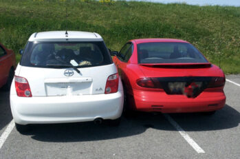 A red car trying to squeeze into a parking spot designed for only one car that is already occupied by a white car.