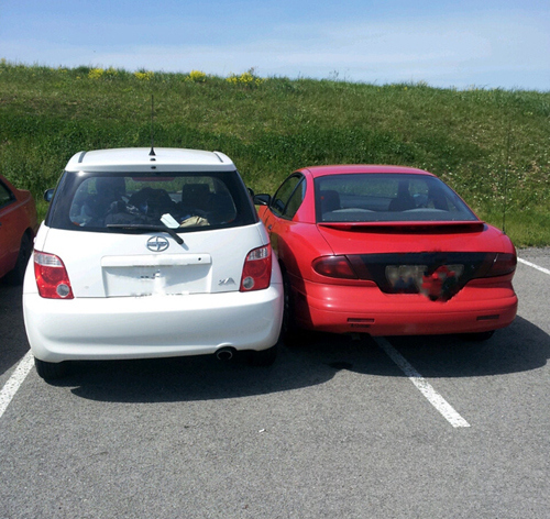 A red car trying to squeeze into a parking spot designed for only one car that is already occupied by a white car.