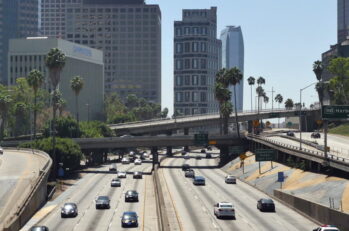 Image of an interstate with a bunch of cars driving on it with buildings in the background.