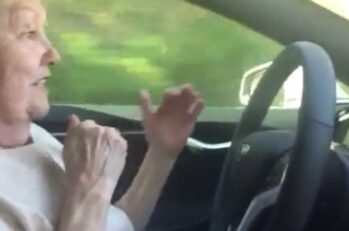 An elderly lady driving a car with her hands off of the steering wheel.