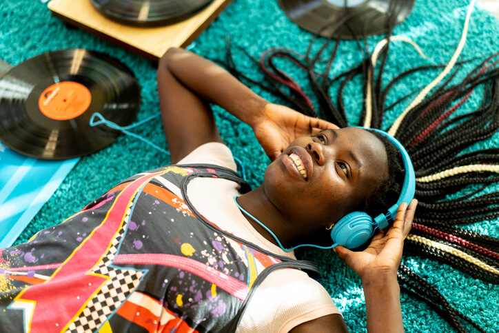An African-American teen is lying on the living room carpet wearing headphones as she listens to some music, surrounded by vinyl records