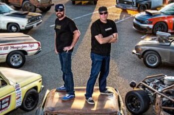 Two guys standing back to back on the hood of a car surrounded by souped up muscle cars.