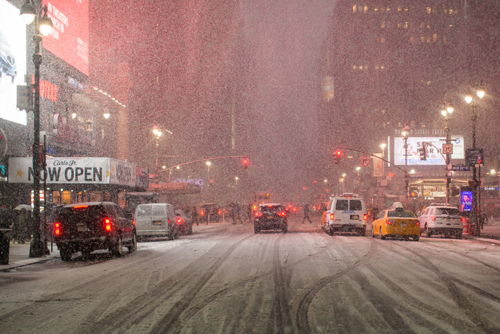 Office workers and tourists are crossing street on a crosswalk in midtown Manhattan. The intersection and the roadway are completely covered by snow. Snow continues to fall and snowflakes are visible flying in the air. The sidewalk is not cleared of snow at all. It's very hard to walk. Winter storm in progress and snow gathers in drifts. High-rise buildings on the left and right. In the distance a skyscraper is in snowy haze. Historical Buildings. Penn station area. Cars parked at the sides of the street are also covered with snow. A group of people stands at the crossroads waiting for a traffic light. Cars drive along the roadway completely covered by snow. Midtown Manhattan. New York. USA